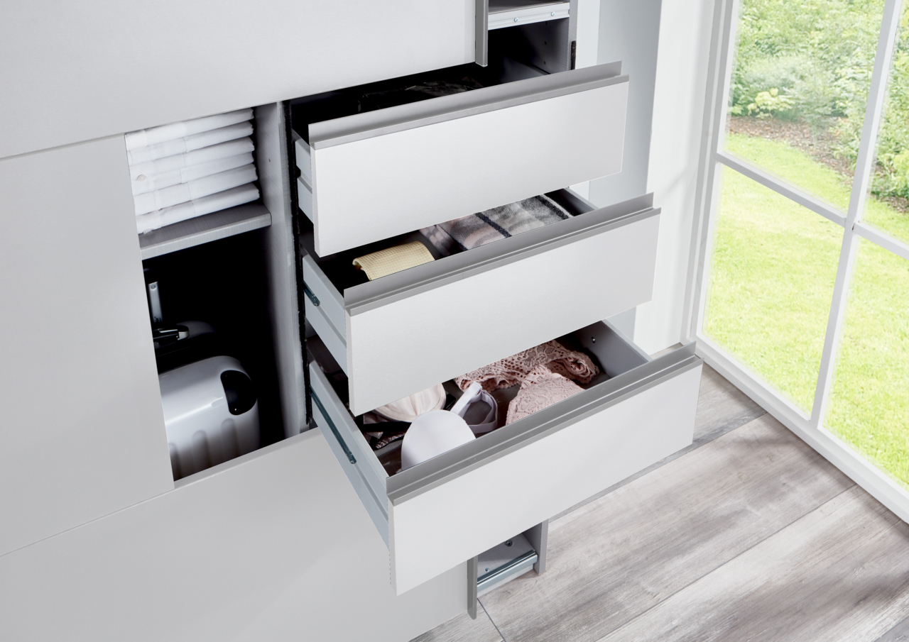 Nahaufnahme eines Schwebetürenschranks mit geöffneten Schubladen, die Kleidung und Accessoires zeigen, neben einem Fenster mit Blick ins Grüne.