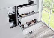 Nahaufnahme eines Schwebetürenschranks mit geöffneten Schubladen, die Kleidung und Accessoires zeigen, neben einem Fenster mit Blick ins Grüne.