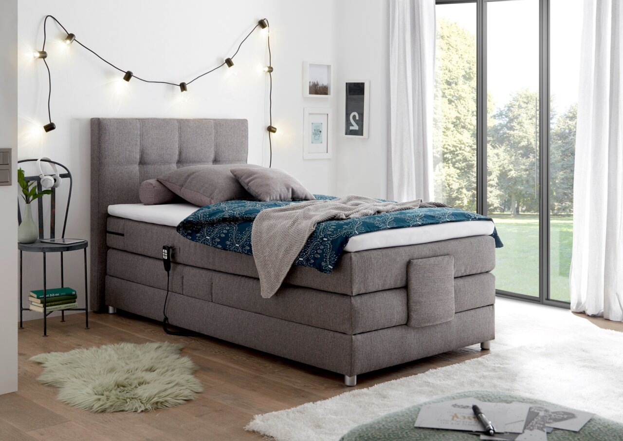 Graues Boxspringbett in einem modernen Schlafzimmer, seitliche Perspektive, mit dekorativen Kissen und Decke, neben einem Fenster mit Blick ins Grüne.