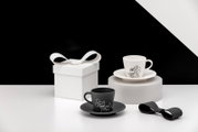 Zwei Espresso-Tassen mit Untertassen in Schwarz und Weiß mit Cartoon-Motiv, neben einer weißen Geschenkbox mit Schleife auf minimalistischem Set; Perspektive: leicht erhöhte Frontansicht.