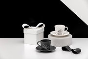 Zwei Espresso-Tassen mit Untertassen in Schwarz und Weiß mit Cartoon-Motiv, neben einer weißen Geschenkbox mit Schleife auf minimalistischem Set; Perspektive: leicht erhöhte Frontansicht.