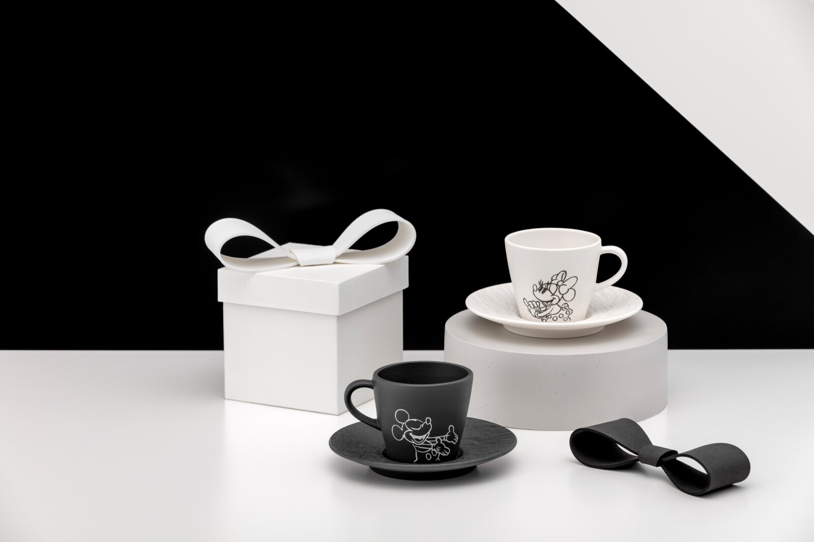 Zwei Espresso-Tassen mit Untertassen in Schwarz und Weiß mit Cartoon-Motiv, neben einer weißen Geschenkbox mit Schleife auf minimalistischem Set; Perspektive: leicht erhöhte Frontansicht.