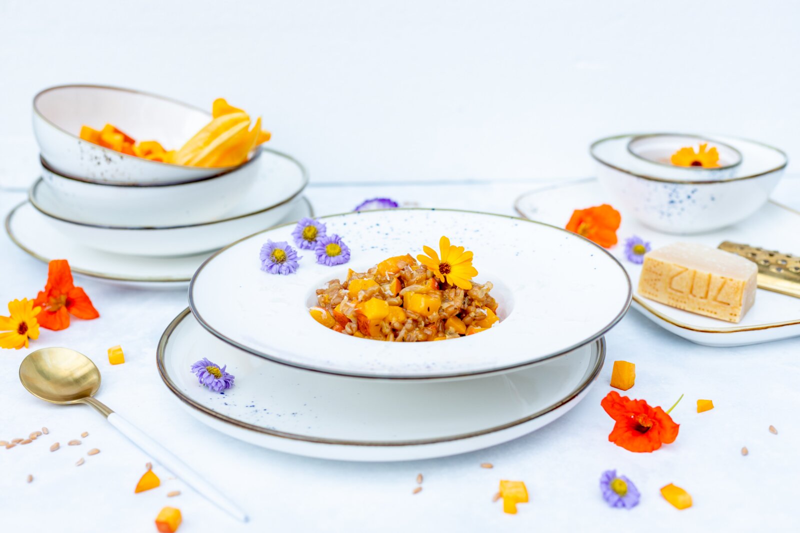 Weiß gesprenkeltes Porzellan-Geschirrset mit tiefem Pastateller, Schalen und Tellern mit braunem Rand, dekorativ angerichtet mit Speise und Blüten, fotografiert in leicht erhöhter Frontansicht.