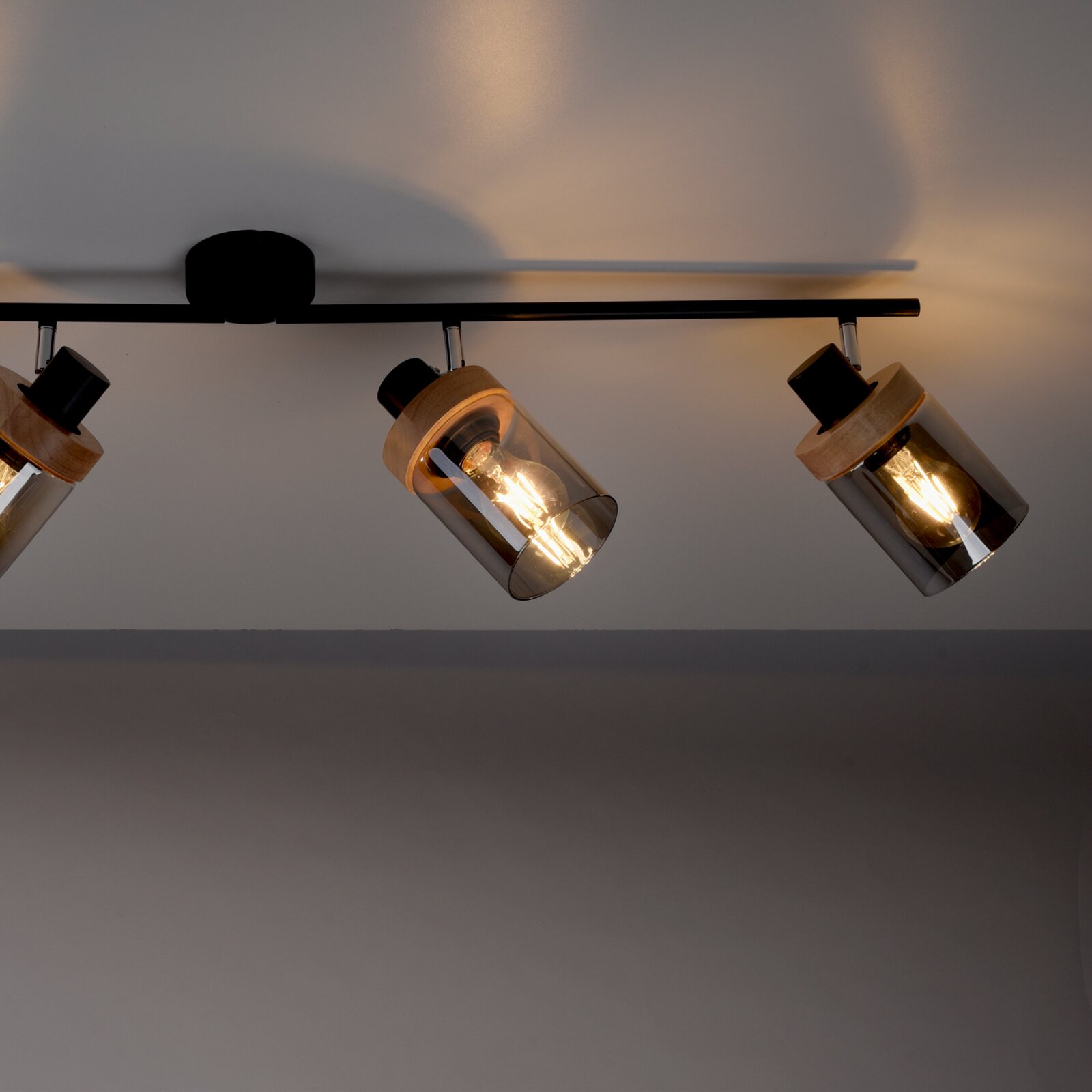 Moderne Deckenleuchte/Spotleiste mit drei verstellbaren Spots, Rauchglas-Zylinderschirmen und Holzdetails, warm leuchtenden Filament-Leuchtmitteln, aus leicht schräger Untersicht fotografiert
