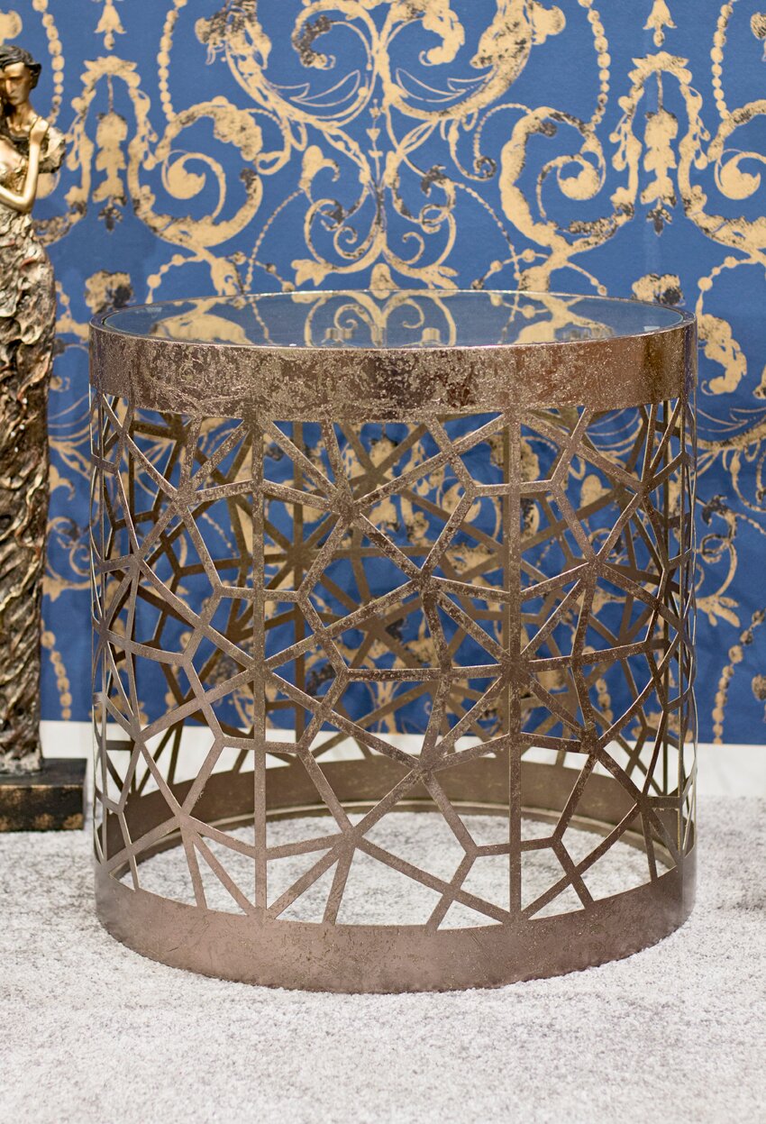 Runder Beistelltisch aus Metall in Antik-Optik mit geometrischem Gittergestell und runder Glasplatte, frontal in leichter Aufsicht fotografiert vor blau-goldener Ornamenttapete.