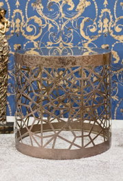 Runder Beistelltisch aus Metall in Antik-Optik mit geometrischem Gittergestell und runder Glasplatte, frontal in leichter Aufsicht fotografiert vor blau-goldener Ornamenttapete.