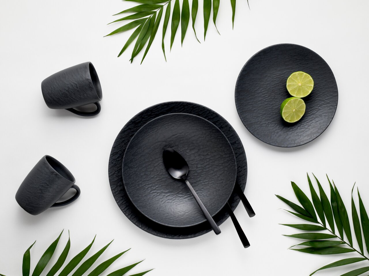 Schwarzes Kombiservice 16-teilig mit Tellern, Tassen und Besteck, aus der Vogelperspektive, dekoriert mit Limetten und grünen Blättern.