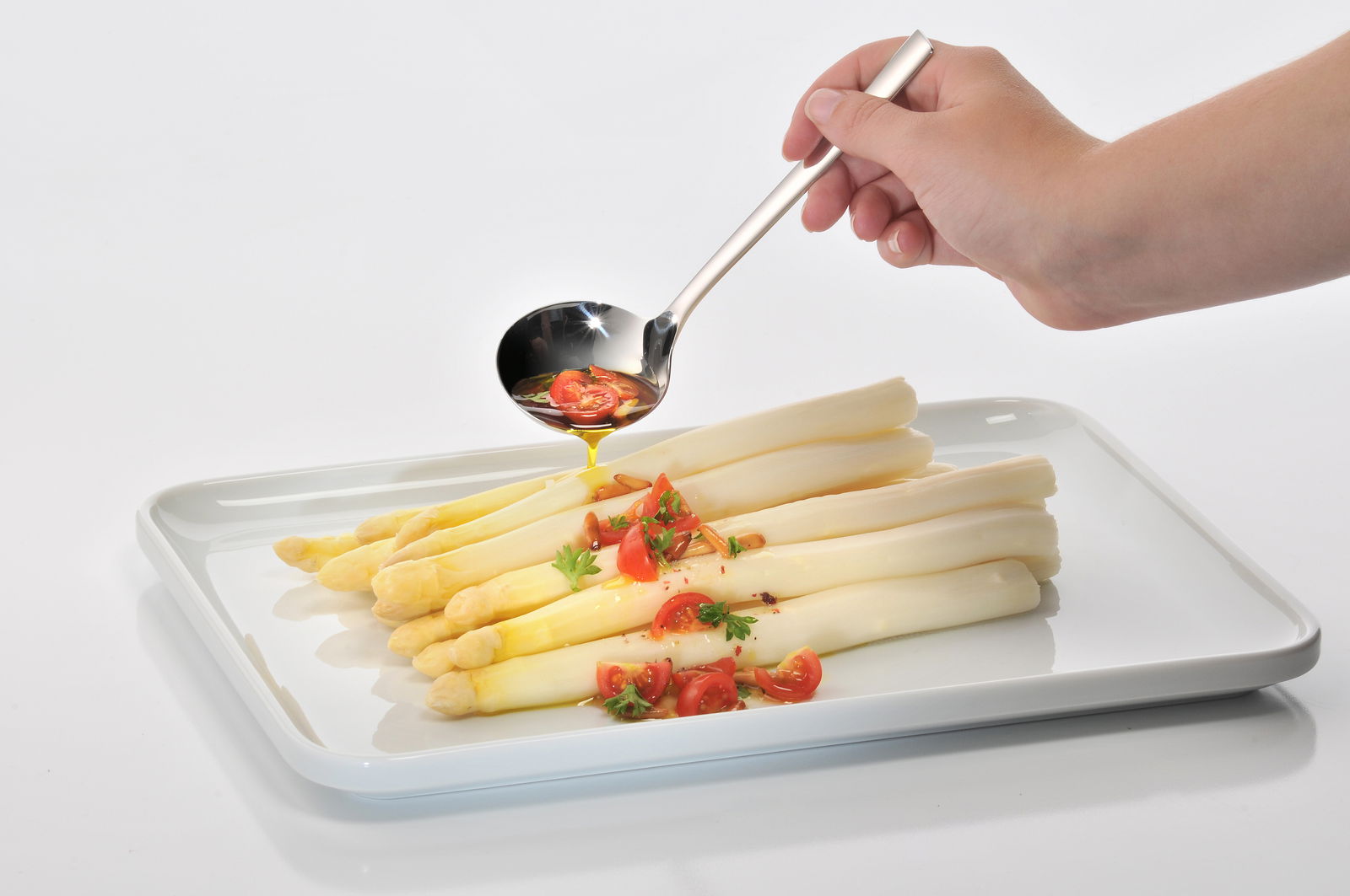 WMF Suppenschöpfer NUOVA Hand hält einen glänzenden Suppenschöpfer über einem Teller mit weißem Spargel und Tomaten, seitliche Perspektive.