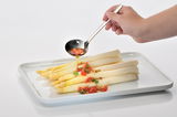 Hand hält einen glänzenden Suppenschöpfer über einem Teller mit weißem Spargel und Tomaten, seitliche Perspektive.