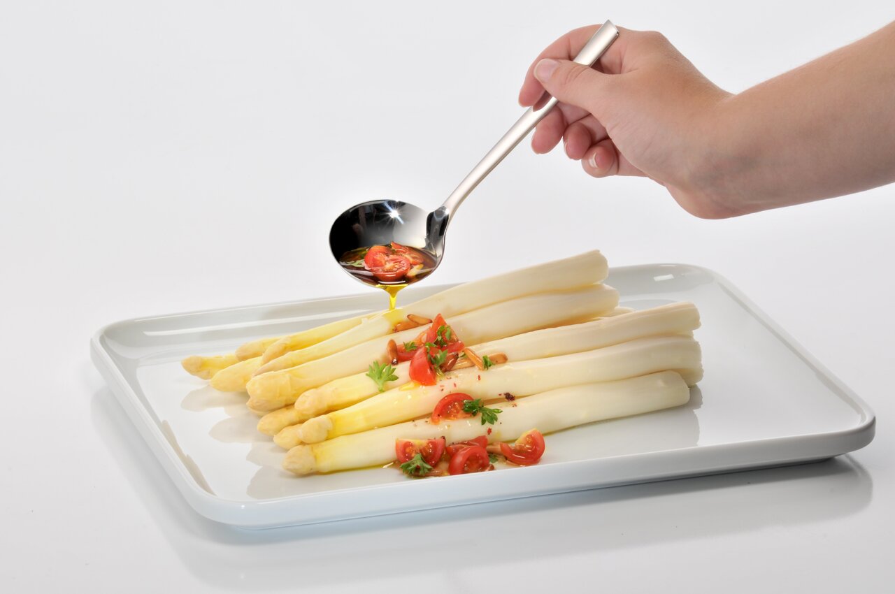 Hand hält einen glänzenden Suppenschöpfer über einem Teller mit weißem Spargel und Tomaten, seitliche Perspektive.