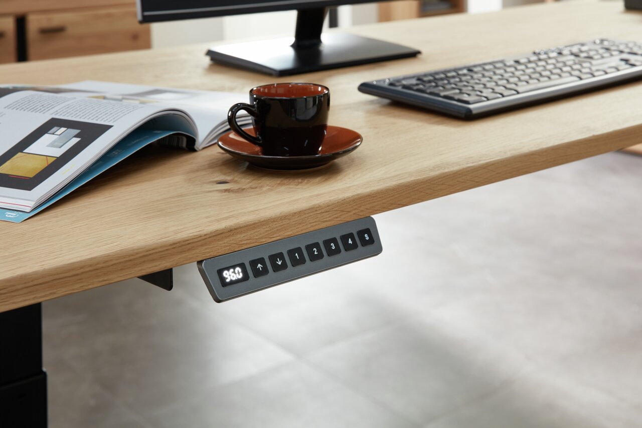 Nahaufnahme eines E-Schreibtischs mit Furnieroberfläche, auf dem eine Tasse, eine Tastatur und ein Magazin liegen. Im Vordergrund ist ein Bedienfeld zur Höhenverstellung sichtbar.