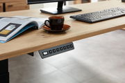 Nahaufnahme eines E-Schreibtischs mit Furnieroberfläche, auf dem eine Tasse, eine Tastatur und ein Magazin liegen. Im Vordergrund ist ein Bedienfeld zur Höhenverstellung sichtbar.