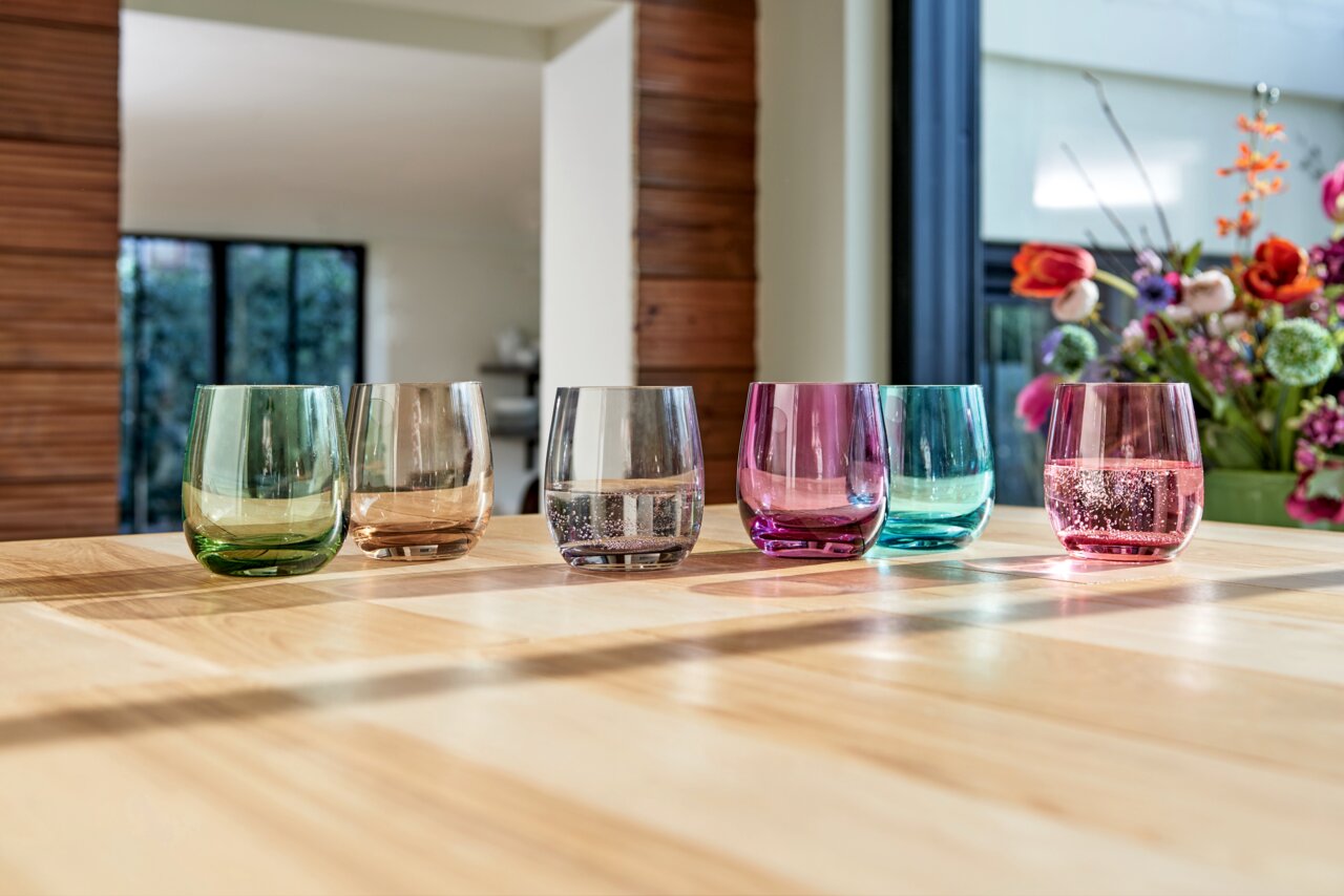 Sechs bunte Trinkgläser aus der Leonardo SORA Kollektion auf einem Holztisch, fotografiert aus einer seitlichen Perspektive mit einem blumigen Hintergrund.