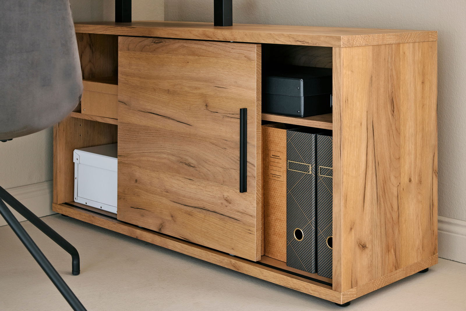 Nahaufnahme eines Holzschranks mit Schiebetür, schwarzen Griffen und mehreren Fächern, gefüllt mit Ordnern und Boxen, von der Seite betrachtet.