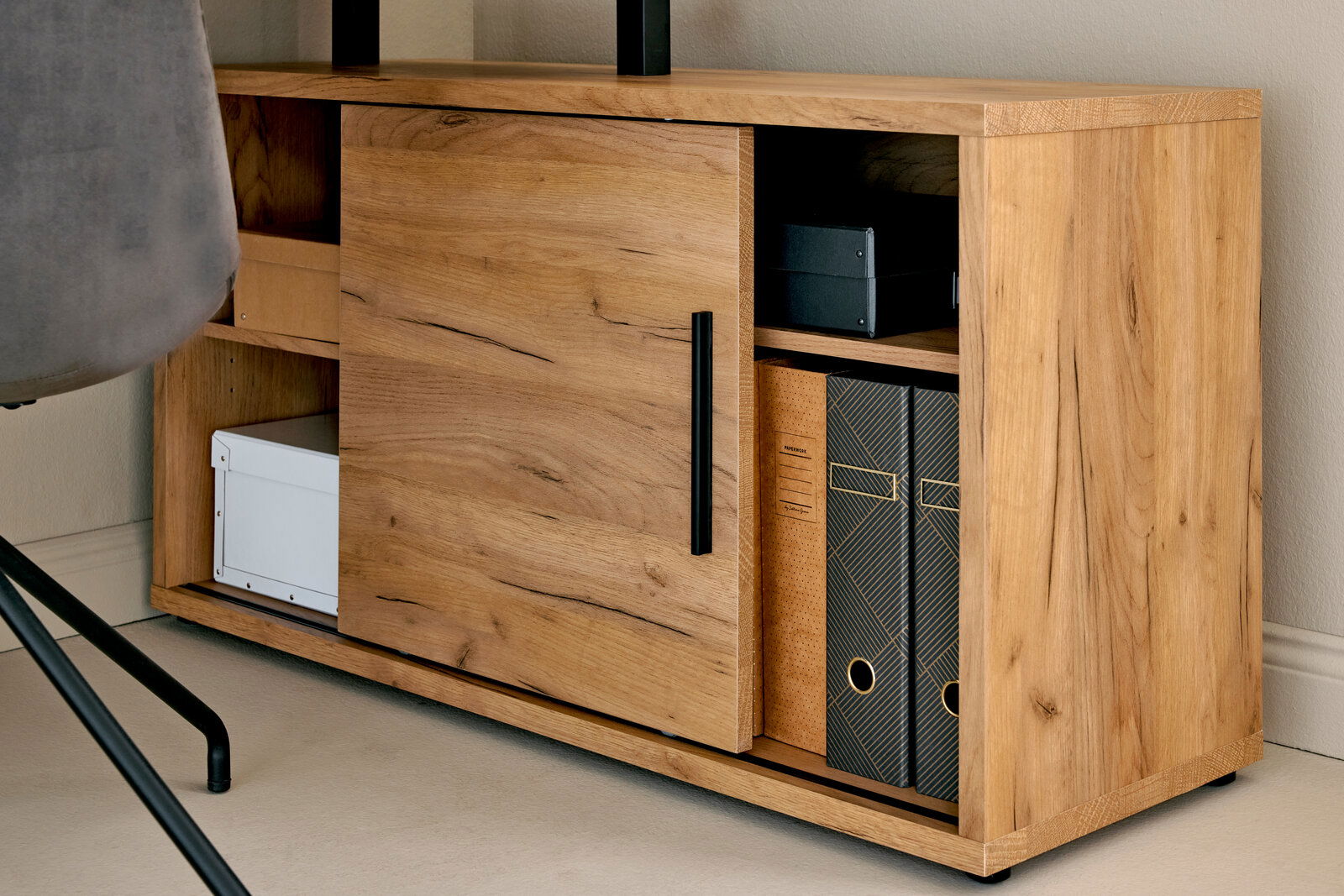 Nahaufnahme eines Holzschranks mit Schiebetür, schwarzen Griffen und mehreren Fächern, gefüllt mit Ordnern und Boxen, von der Seite betrachtet.