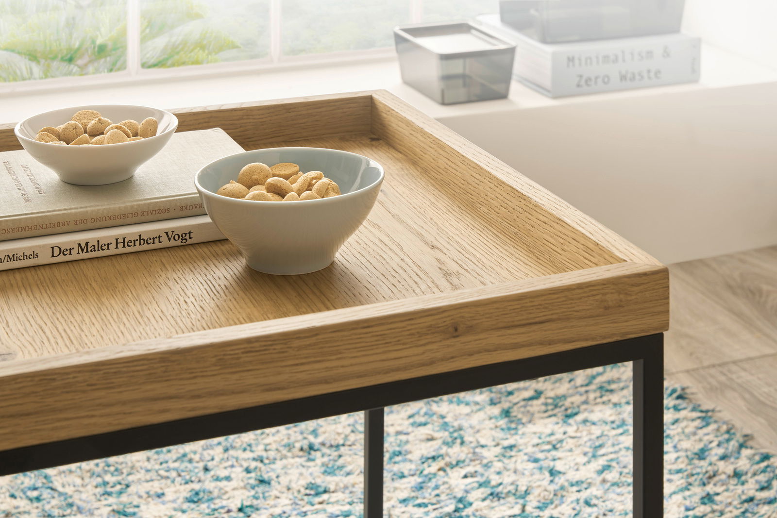 Nahaufnahme eines Satztisches mit Holzoberfläche und schwarzem Metallgestell, darauf zwei Schalen mit Keksen und ein Buch, im Hintergrund ein Fenster mit Blick ins Grüne.