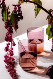 Zwei rosafarbene Longdrinkbecher mit einem Glasstrohhalm, umgeben von dekorativen lila Blüten, aus der Seitenperspektive fotografiert.