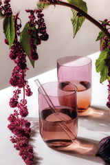 Zwei rosafarbene Longdrinkbecher mit einem Glasstrohhalm, umgeben von dekorativen lila Blüten, aus der Seitenperspektive fotografiert.