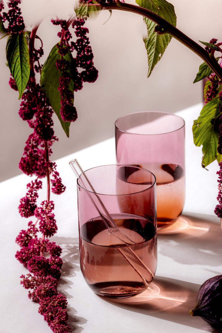 Zwei rosafarbene Longdrinkbecher mit einem Glasstrohhalm, umgeben von dekorativen lila Blüten, aus der Seitenperspektive fotografiert.