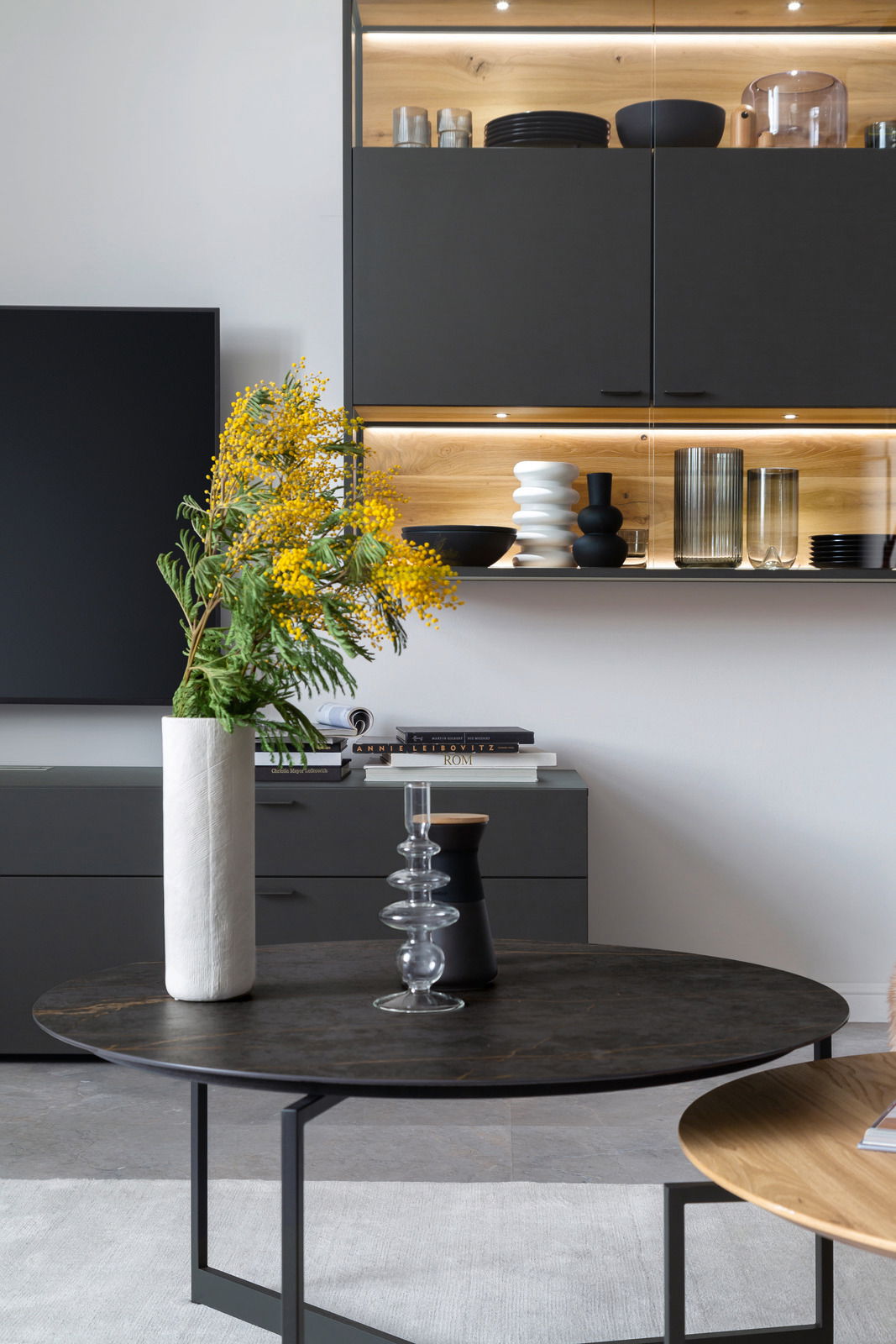 Moderner Couchtisch mit dunkler runder Tischplatte und schlanken Metallbeinen, dekoriert mit einer hohen Vase mit gelben Blumen, aus der Perspektive von vorne.