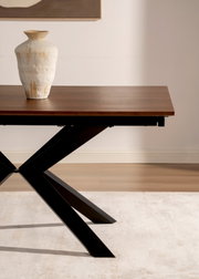 Moderne eettafel met donker houten blad en zwart metalen X-poot, gefotografeerd in driekwart zijaanzicht; op het blad staat een beige keramische vaas in een minimalistisch interieur.