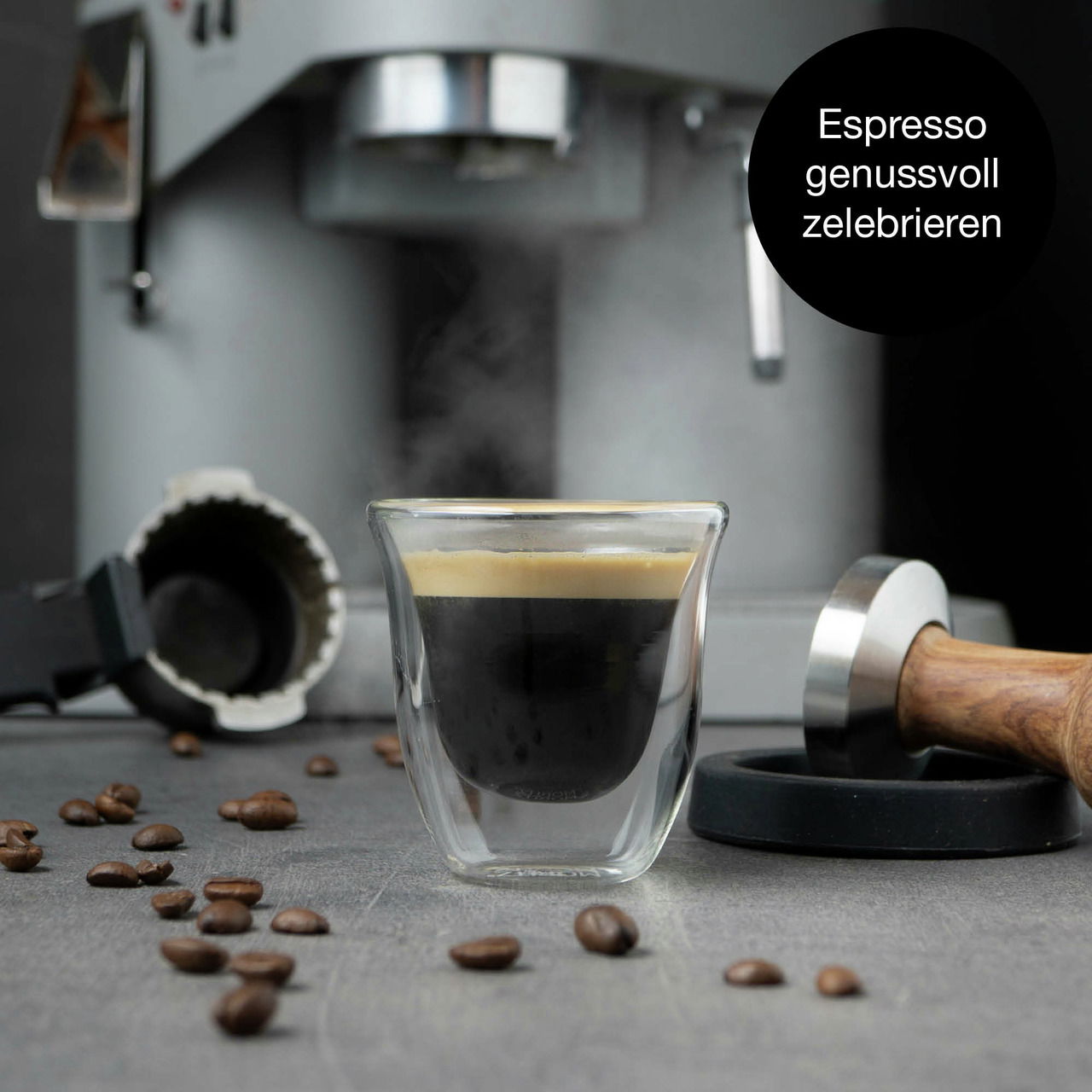 Nahaufnahme eines frisch gebrühten Espressos in einem doppelwandigen Glas vor einer Espressomaschine, umgeben von Kaffeebohnen und einem Tamper.