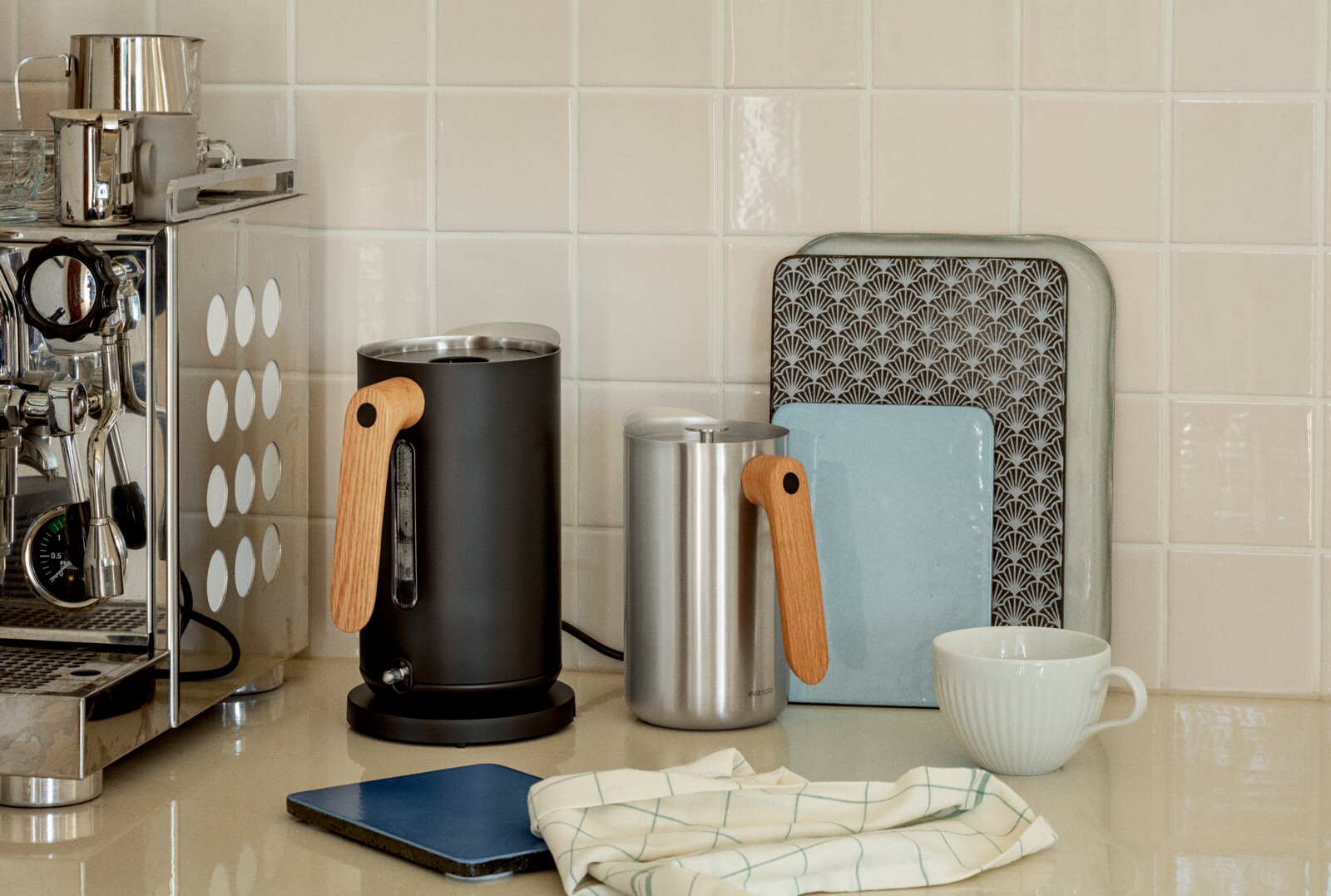 Eva Solo Wasserkocher NORDIC KITCHEN Zwei moderne Wasserkocher mit Holzgriff (schwarz und Edelstahl) auf einer Küchenarbeitsplatte neben Espressomaschine und Tasse, frontale Perspektive auf Augenhöhe.