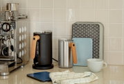 Keukenaanrecht met twee elektrische waterkokers (zwart en roestvrij staal) met houten handgrepen naast een espressoapparaat, snijplanken en een witte kop, gefotografeerd vanuit een frontaal licht schuin zijaanzicht.