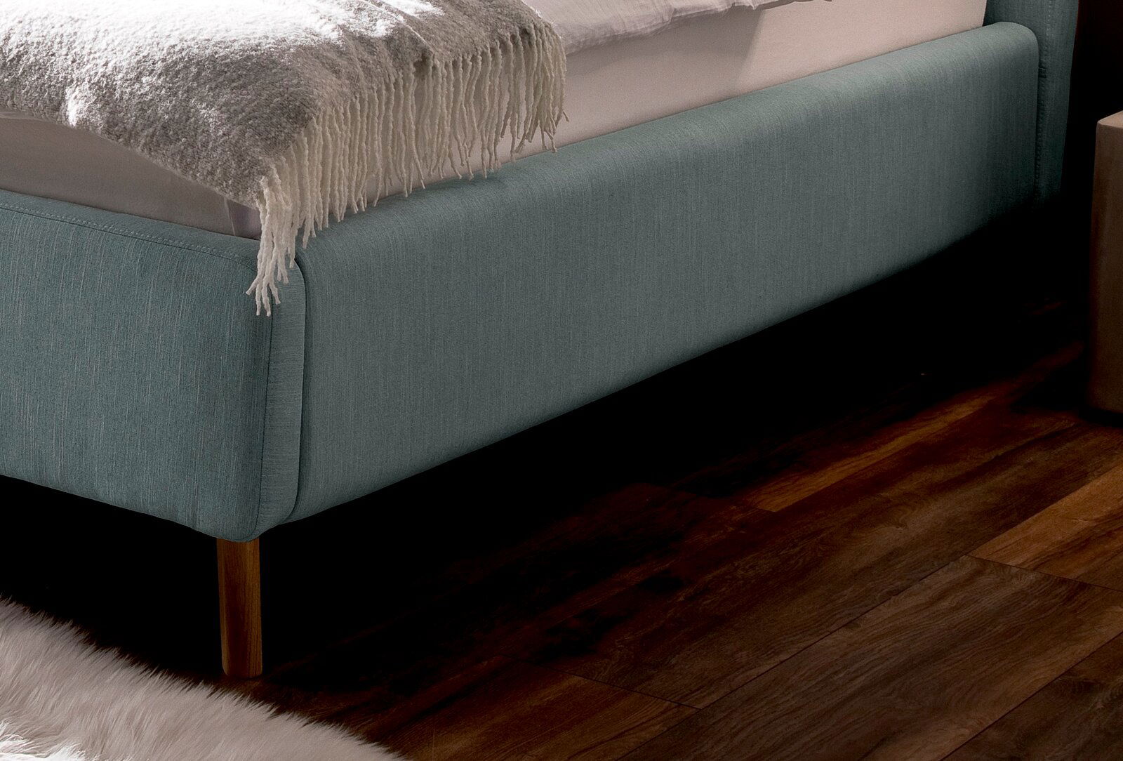 Nahaufnahme eines Polsterbetts in hellblauem Stoff mit Holzfüßen, seitliche Perspektive.