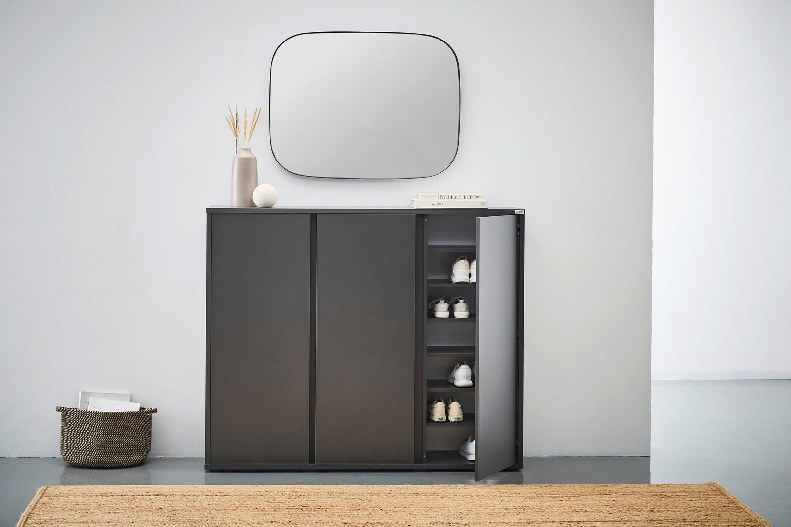 Moderner schwarzer Schuhschrank mit geöffneter Tür, der von vorne gezeigt wird. Im Inneren sind mehrere Paar Schuhe sichtbar. Der Schrank steht in einem minimalistischen Raum mit einem Spiegel darüber und Dekorationen darauf.
