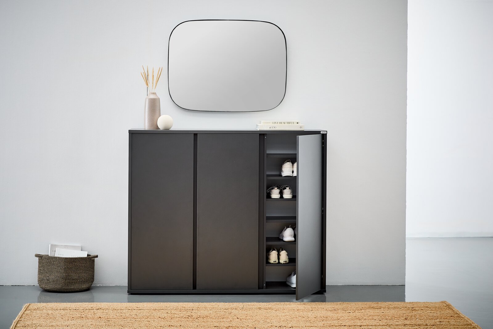 Moderner schwarzer Schuhschrank mit geöffneter Tür, der von vorne gezeigt wird. Im Inneren sind mehrere Paar Schuhe sichtbar. Der Schrank steht in einem minimalistischen Raum mit einem Spiegel darüber und Dekorationen darauf.
