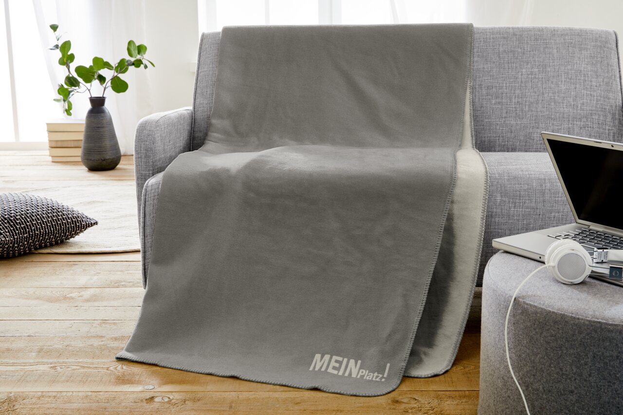 Graue Jacquard-Decke 150x200cm auf einem grauen Sofa, mit der Aufschrift 'MEIN Platz!' in einem modernen Wohnzimmer.