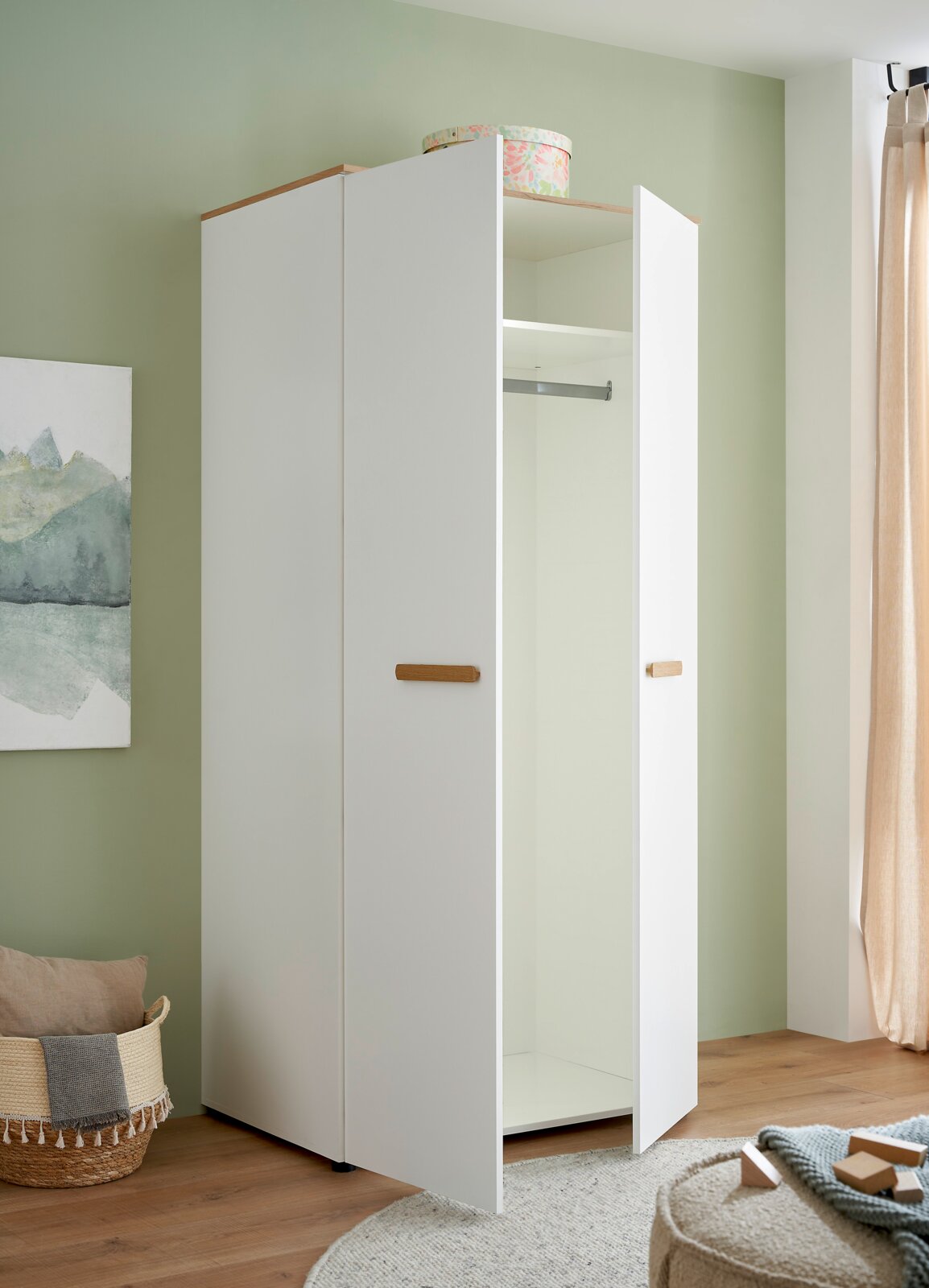 Weißer zweitüriger Kleiderschrank mit Holzgriffen und Holz-Deckplatte, eine Tür geöffnet mit Kleiderstange und Ablagefach sichtbar, Innenansicht aus leicht schräger Frontperspektive im hellen Zimmer
