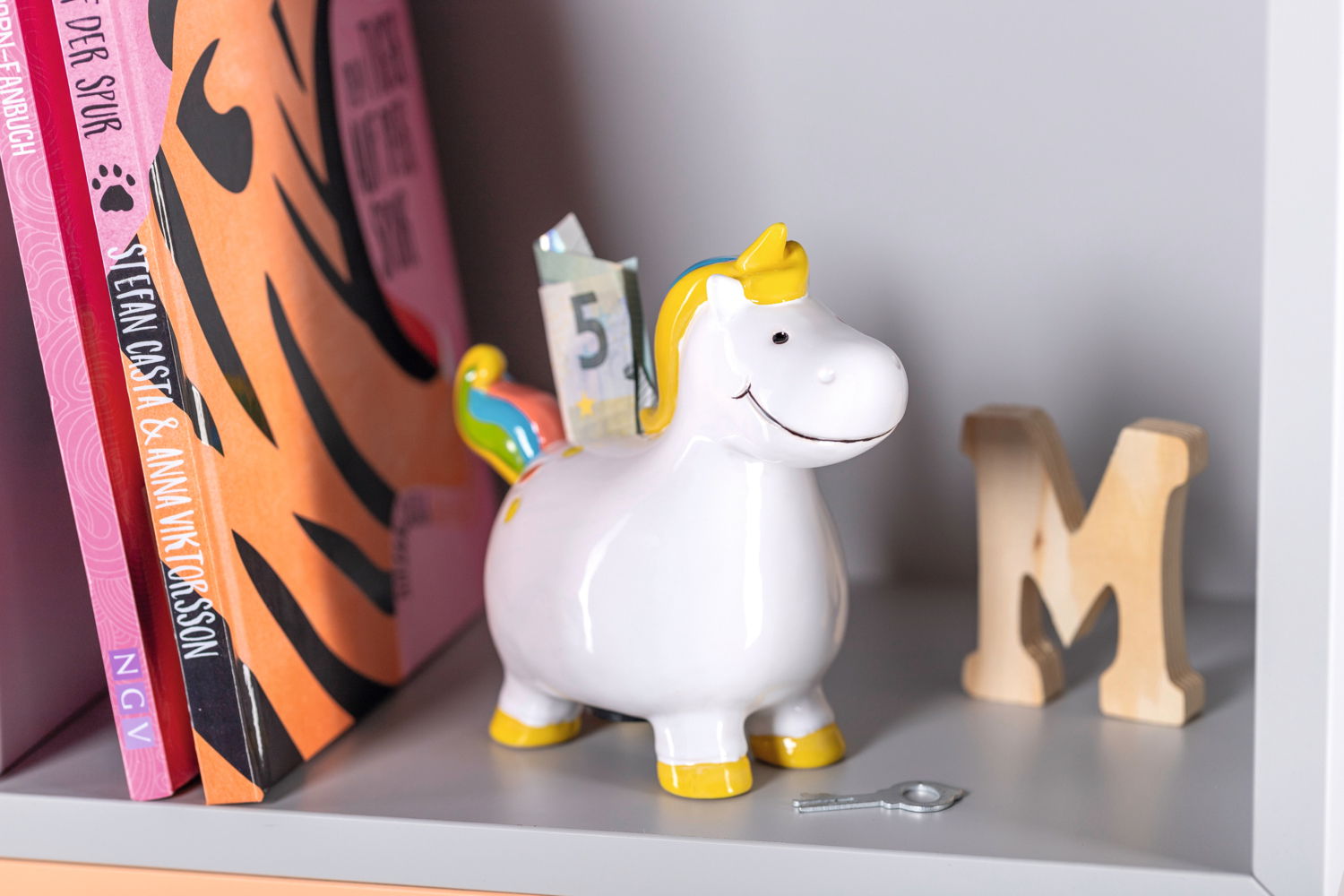 Leonardo Spardose Einhorn BAMBINI Spardose in Form eines Einhorns mit Regenbogenmähne und gelben Hufen, seitliche Perspektive, auf einem Regal neben Büchern und einem Holzbuchstaben.