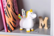 Spardose in Form eines Einhorns mit Regenbogenmähne und gelben Hufen, seitliche Perspektive, auf einem Regal neben Büchern und einem Holzbuchstaben.