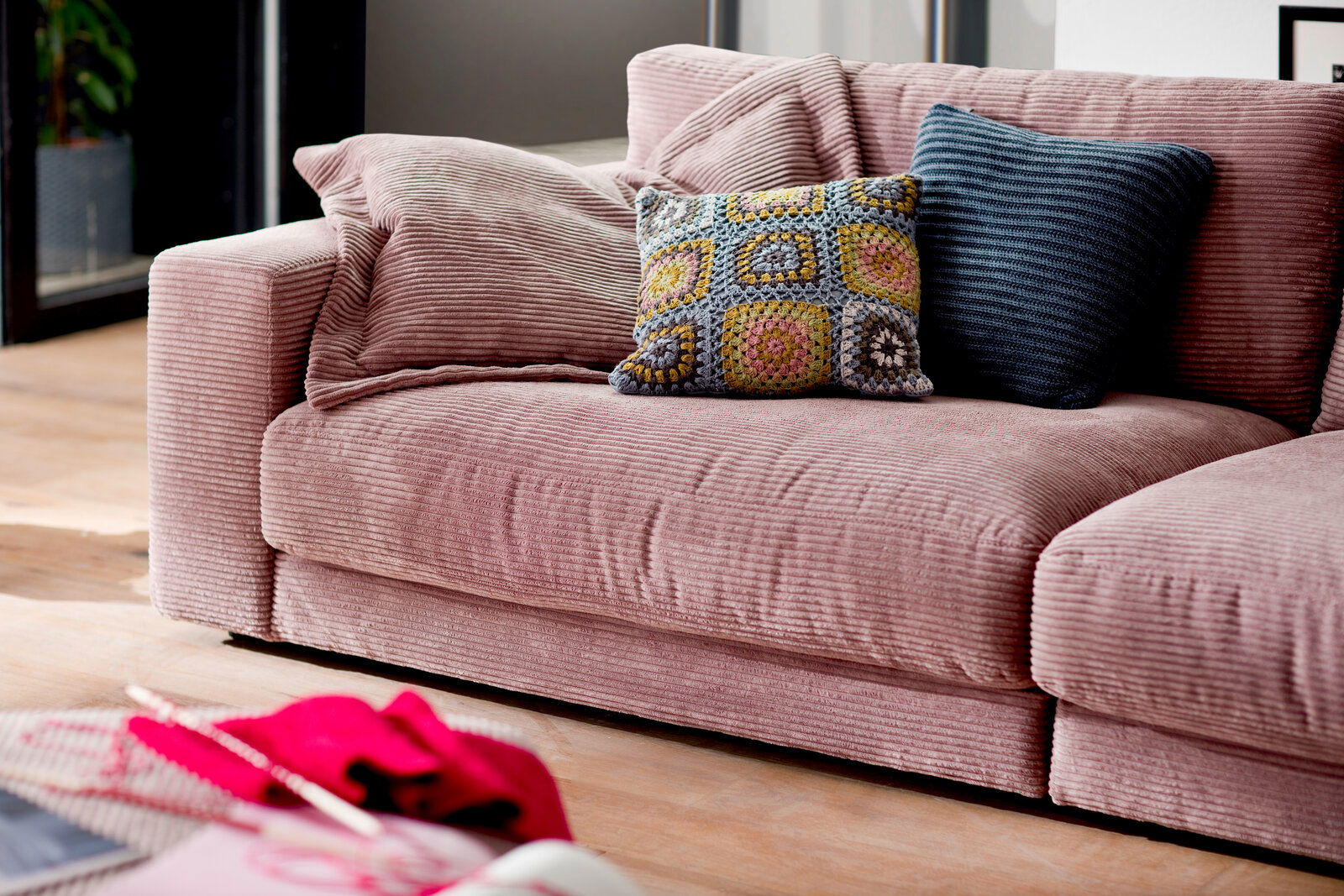 Nahaufnahme eines rosafarbenen 2er-Sofas mit geripptem Stoff, dekoriert mit zwei Kissen, eines in Häkeloptik und eines in Dunkelblau.