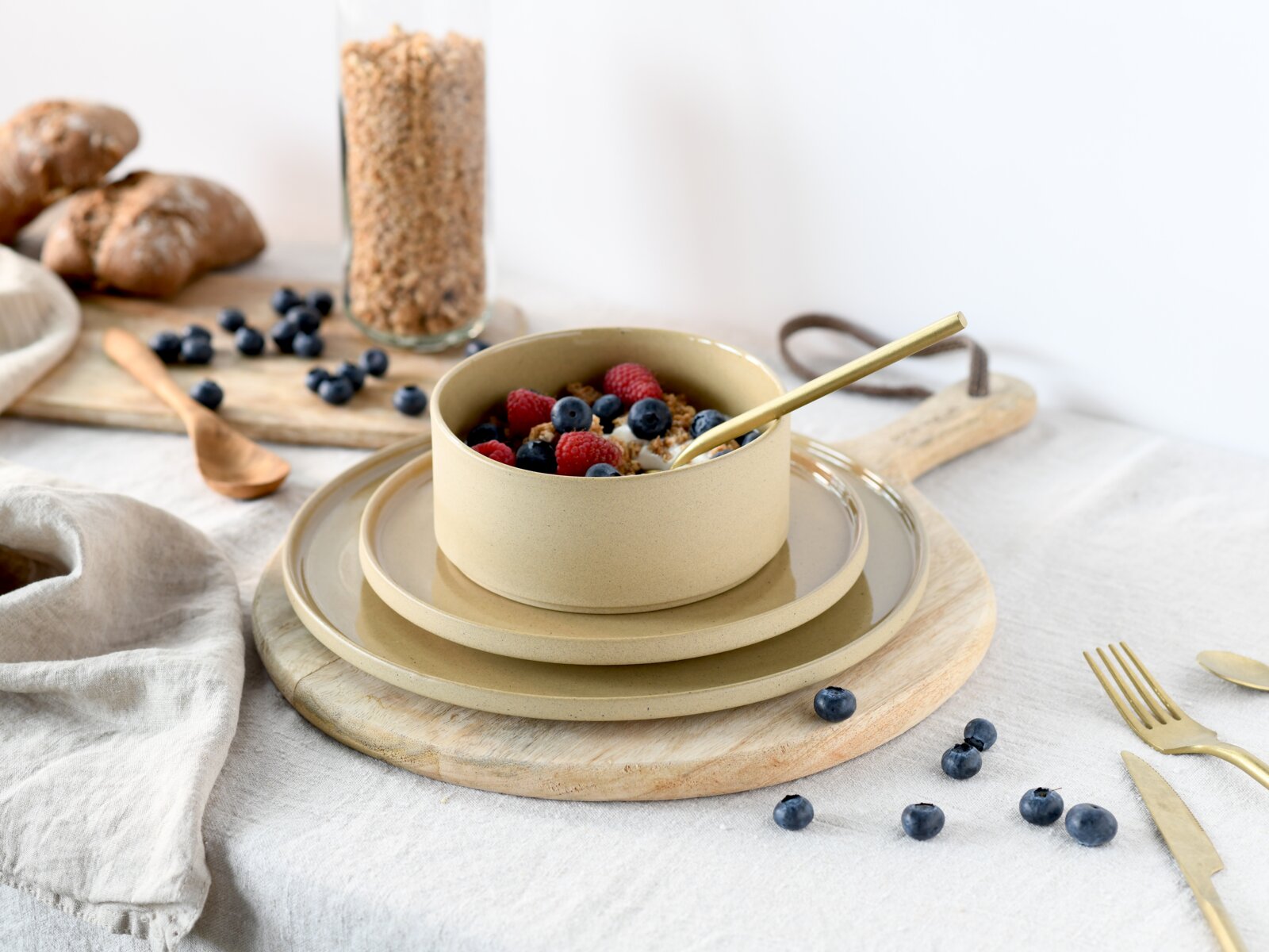 Beige Keramikschale mit Müsli und Beeren auf passenden Tellern, goldfarbener Löffel; Perspektive: leicht von oben in schräger Frontansicht auf einem Holzteller arrangiert.