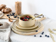 Beige Keramikschale mit Müsli und Beeren auf passenden Tellern, goldfarbener Löffel; Perspektive: leicht von oben in schräger Frontansicht auf einem Holzteller arrangiert.