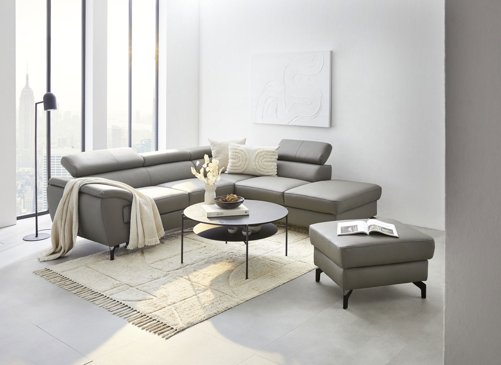 Moderne Eckgarnitur in einem hellen Wohnzimmer mit großen Fenstern, grauem Lederbezug und dekorativen Kissen, aus der Perspektive von vorne links.