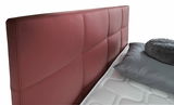 Nahaufnahme eines Boxspringbetts mit rotem, gestepptem Kopfteil und grauen Kissen, seitliche Perspektive.