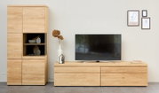 Moderne Wohnwand aus hellem Naturholz mit hohem Vitrinenschrank und breitem TV-Lowboard, frontal in einem stilvoll dekorierten Wohnzimmer dargestellt.