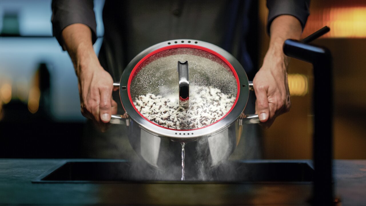 WMF braadpan FUNCTION 4 Een braadpan met deksel, 24 cm, van roestvrij staal, wordt met twee handen boven een gootsteen vastgehouden terwijl het water wordt afgegoten. Vooraanzicht.