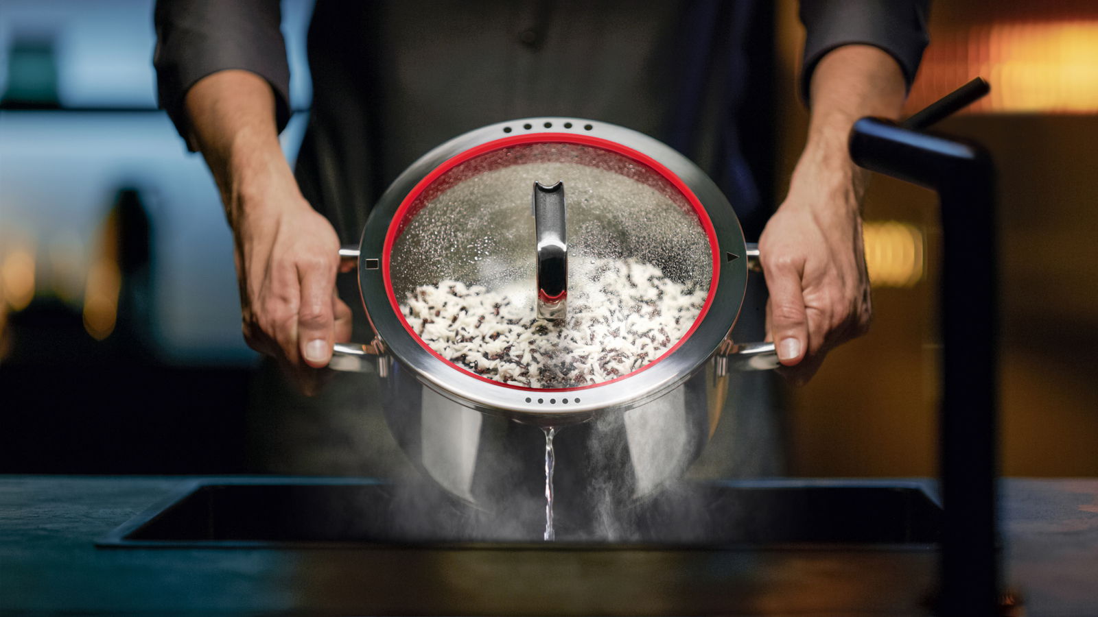 WMF Bratentopf Bratentopf mit Deckel, 24 cm, aus Edelstahl, wird von zwei Händen über einem Waschbecken gehalten, während Wasser abgelassen wird. Perspektive von vorne.