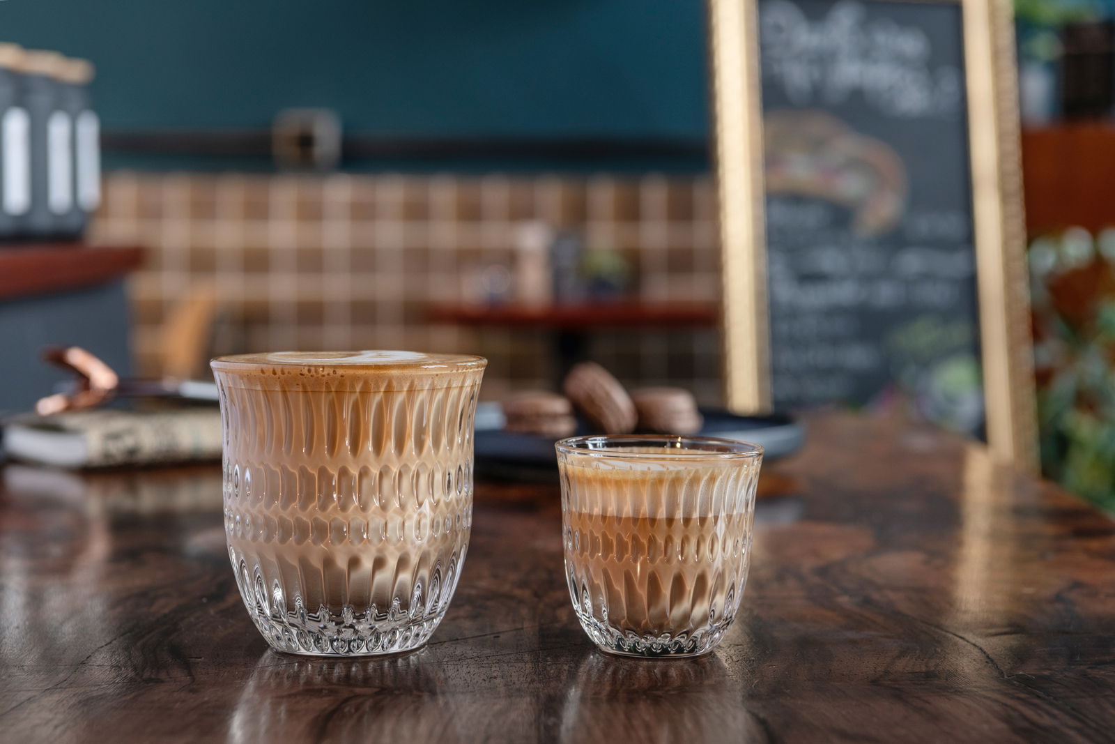 Zwei Cappuccino-Gläser auf einem Holztisch, im Hintergrund ein Café mit unscharfer Tafel. Perspektive von vorne.
