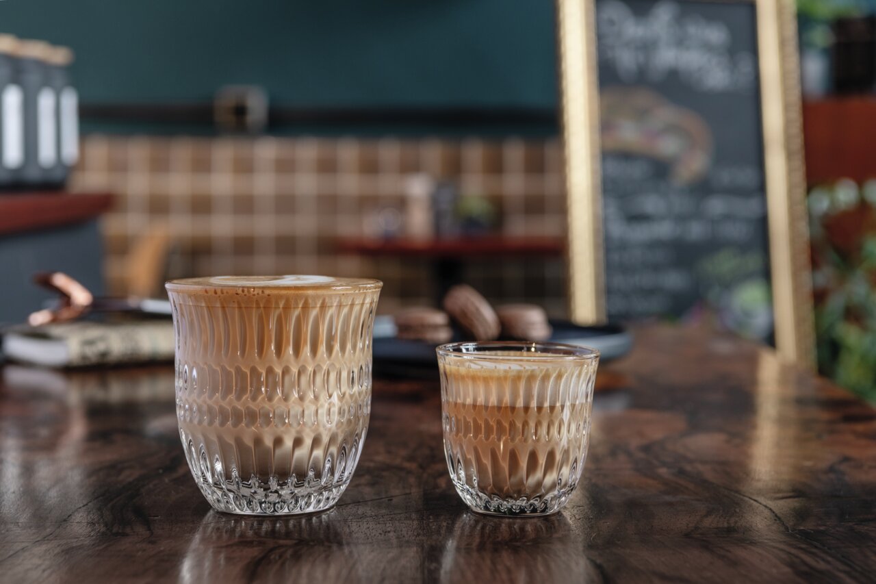 Twee cappuccinoglazen op een houten tafel, op de achtergrond een café met een wazig bord. Vooruitkijkend perspectief.
