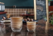 Twee cappuccinoglazen op een houten tafel, op de achtergrond een café met een wazig bord. Vooruitkijkend perspectief.