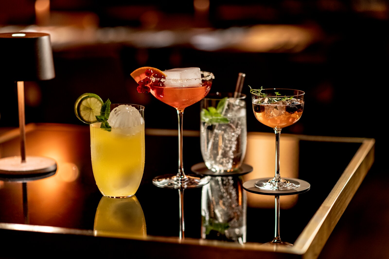 Vier elegante Cocktails auf einem spiegelnden Tablett in einer Bar, fotografiert in frontaler Tischperspektive mit leicht seitlichem Blickwinkel gelber Drink mit Limettenscheibe und Minze, roséfarbener Cocktail mit Zuckerrand und Beeren, Highball mit Eis