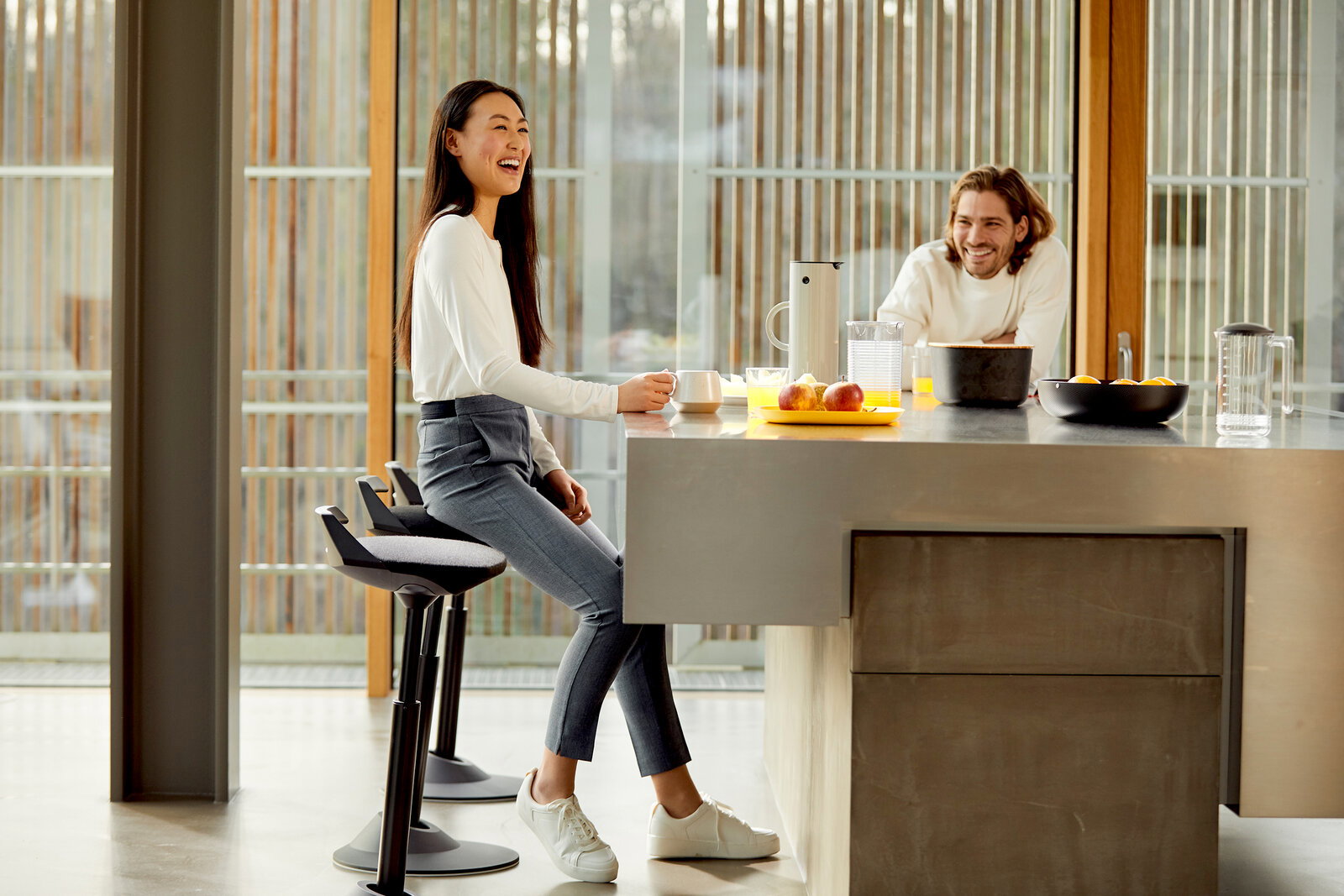 Eine Frau sitzt auf einer Aeris Sitz-/Stehhilfe Muvman an einem Küchentresen, während ein Mann gegenüber steht und lächelt. Die Perspektive zeigt die Seite der Sitz-/Stehhilfe.