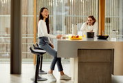 Eine Frau sitzt auf einer Aeris Sitz-/Stehhilfe Muvman an einem Küchentresen, während ein Mann gegenüber steht und lächelt. Die Perspektive zeigt die Seite der Sitz-/Stehhilfe.