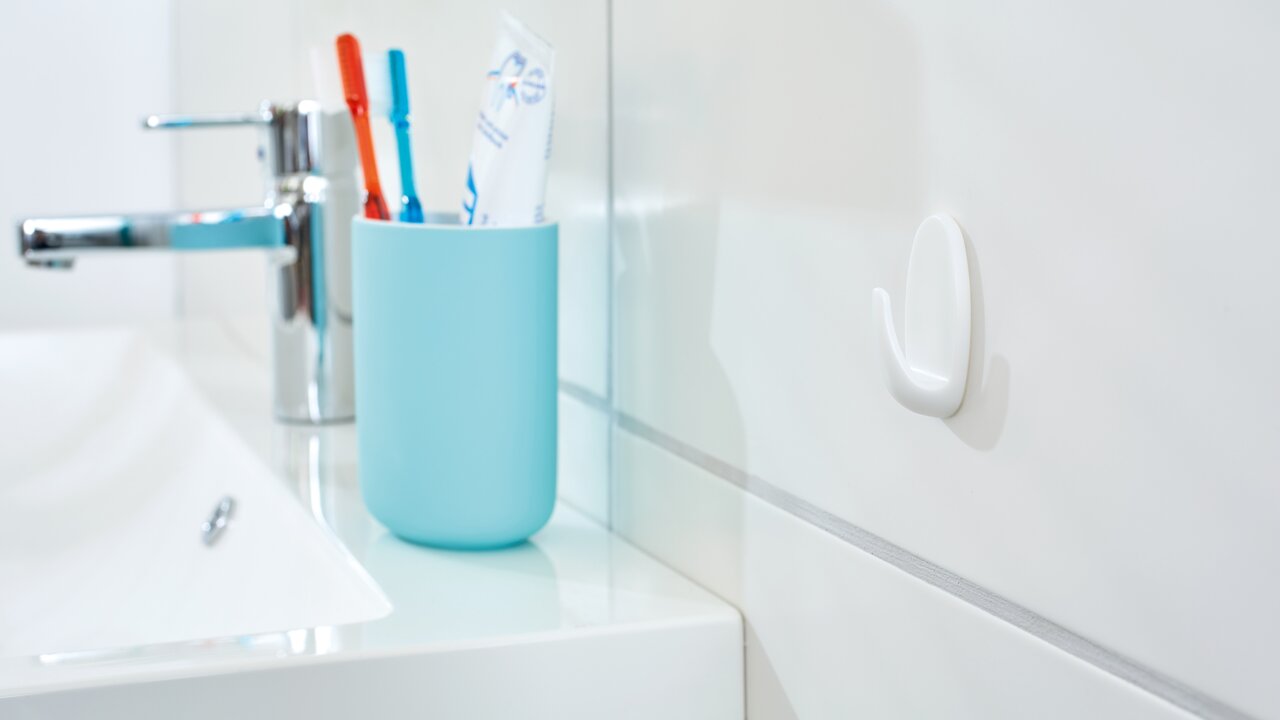 Tesa Powerstrips Haken 3-tlg. Weißer tesa Powerstrips Haken Small an einer Badezimmerwand, seitliche Perspektive mit Waschbecken und Zahnbürstenhalter im Hintergrund.